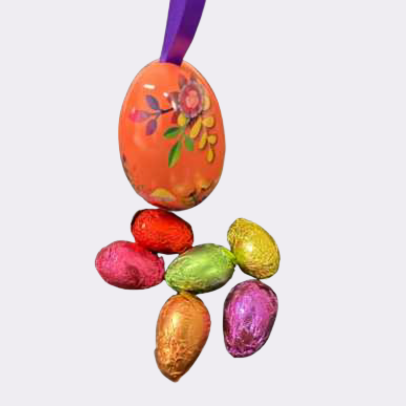 Une décoration de Pâques en forme d'œuf suspendu, colorée en orange vif avec des motifs floraux et une attache en ruban violet. L'œuf est accompagné de six petits œufs pralinés emballés dans du papier métallisé de différentes couleurs (rouge, rose, doré, vert et violet).