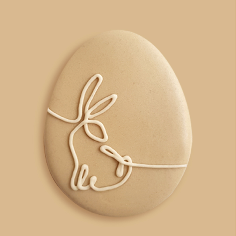 Biscuit de Pâques en forme d'œuf, orné d'un dessin de lapin en glaçage blanc, posé sur un fond beige doux.