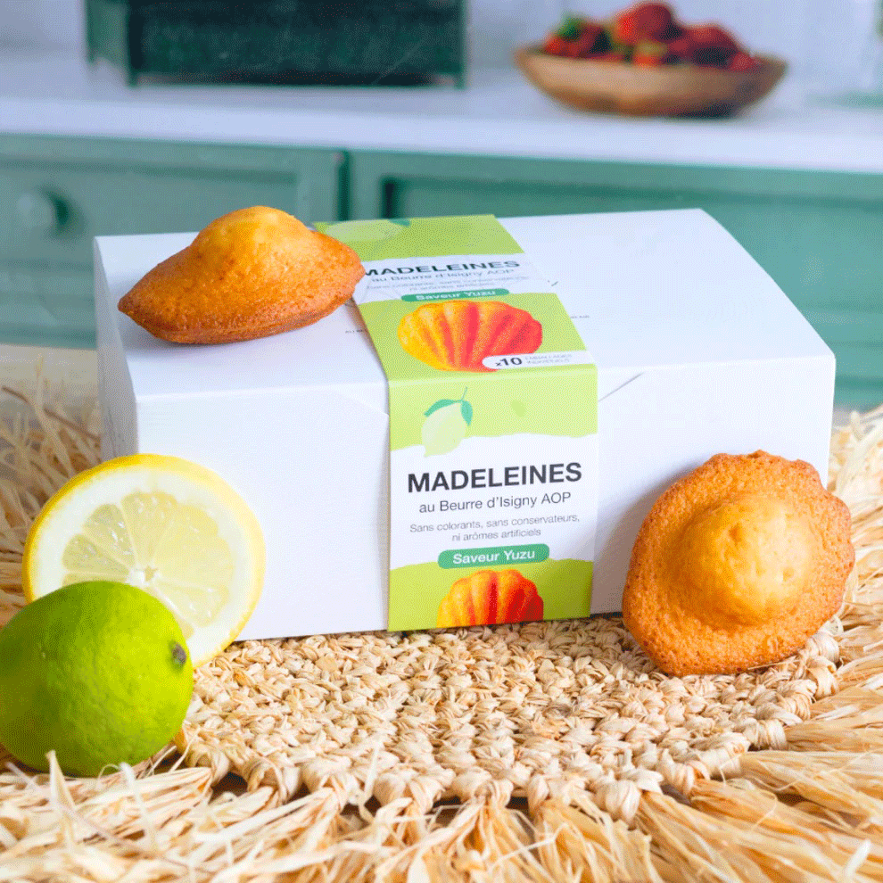 Boîte de madeleines au beurre d'Isigny AOP saveur yuzu, entourée de madeleines, d’un citron et d’un citron vert.