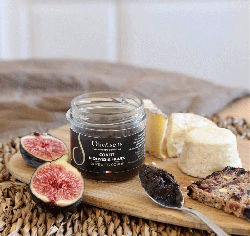 Pot en verre de « Confit d’olives & figues » Oliv&sens ouvert avec une cuillère, accompagné de fromages, de figues fraîches et d’une tranche de pain sur une planche en bois. 