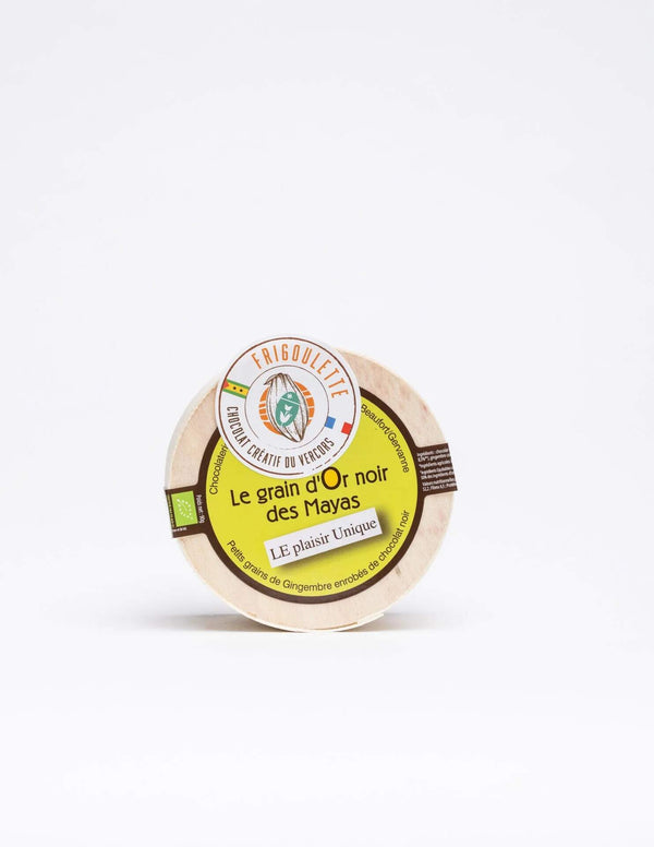 Pot rond en bois avec une étiquette colorée indiquant "Le grain d'Or noir des Mayas"  Frigoulette. L'étiquette mentionne que le produit contient des grains de gingembre enrobés de chocolat noir, et arbore un drapeau de São Tomé-et-Principe.