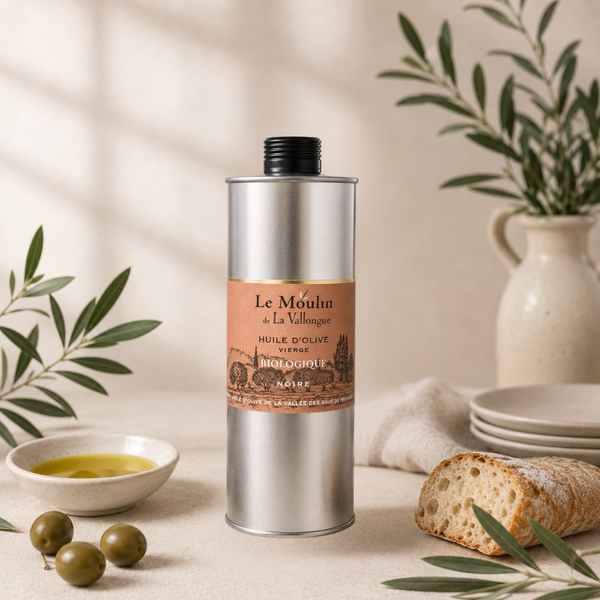 Huile d’olive vierge biologique noire en bidon métallique, présentée avec olives vertes, pain rustique et branches d’olivier sur fond lumineux.