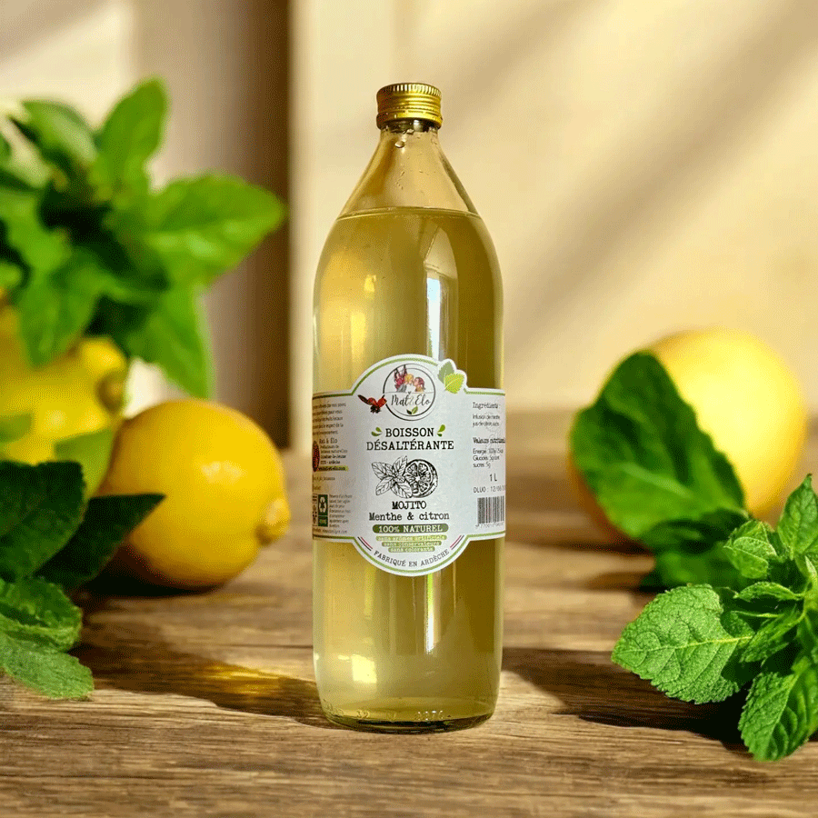 Bouteille en verre d’infusion glacée mojito à la menthe et au citron, de marque Mate&Elo. L’étiquette blanche indique ‘Boisson désaltérante, 100% naturelle, fabriquée en Ardèche’. Le liquide est d’une teinte jaune pâle. En arrière-plan, plusieurs citrons jaunes et des feuilles de menthe verte sont disposés sur une surface en bois naturel.