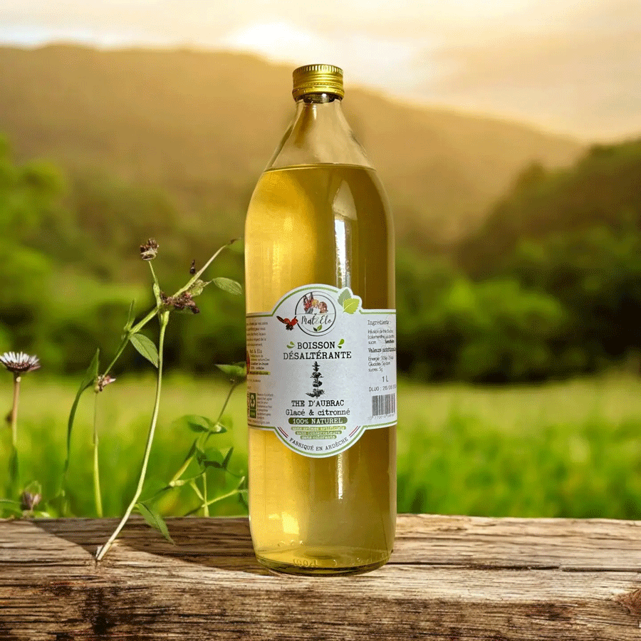 Bouteille en verre d’infusion glacée thé d’Aubrac citronné, de marque Nat&Co. L’étiquette blanche indique ‘Boisson désaltérante, 100% naturelle, fabriquée en Ardèche’. Le liquide est d’une teinte dorée claire. La bouteille est posée sur une surface en bois avec en arrière-plan un paysage naturel verdoyant et des collines au coucher du soleil.