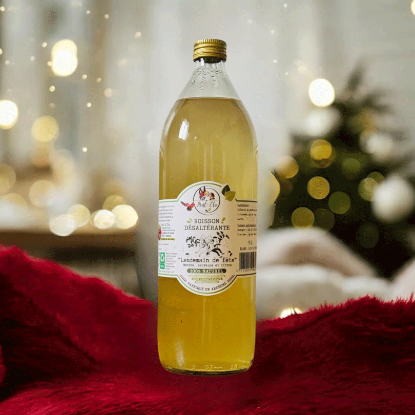 Bouteille en verre d’infusion glacée ‘Lendemain de fête’ à la menthe, verveine et citron, de marque Mat&Elo. L’étiquette blanche mentionne ‘Boisson désaltérante, 100% naturel, fabriquée en Ardèche’, avec une illustration de plantes aromatiques. Le liquide est d’une teinte jaune doré. La bouteille est posée sur un tissu rouge en velours, avec en arrière-plan un sapin de Noël illuminé et des lumières dorées floues.