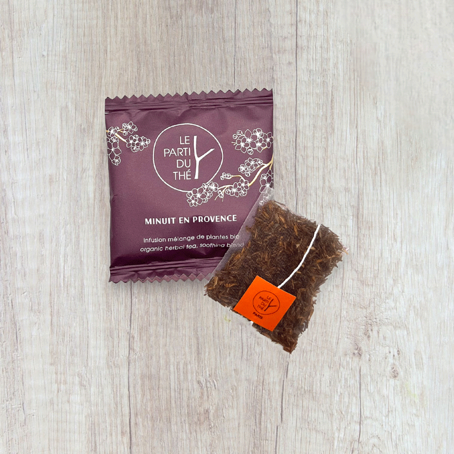 Sachet d'infusion "Minuit en Provence" de la marque Le Parti du Thé, emballage violet décoré de fleurs blanches. À côté, un sachet de thé en mousseline rempli d’herbes sèches, avec une étiquette orange portant le logo ‘Le Parti du Thé, Paris’. Le tout est posé sur une table en bois clair.