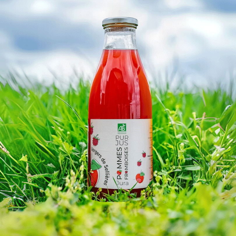 Bouteille de 1 litre de pur jus pommes framboises bio posée au milieu d’une herbe verte et fraîche. L’étiquette blanche affiche la mention "AB" (Agriculture Biologique), des illustrations de framboises et de pommes, ainsi que le nom du producteur "Vergers de Sellières". Le jus est d’un rouge éclatant.