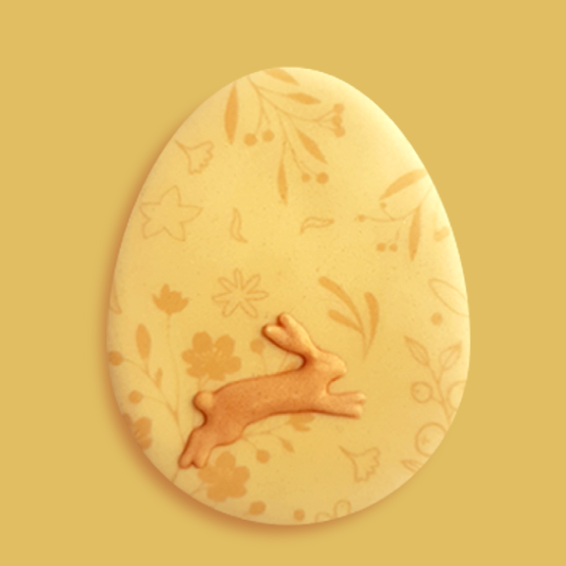 Biscuit de Pâques en forme d'œuf jaune, orné d'un lapin bondissant en relief et de motifs floraux légers. 