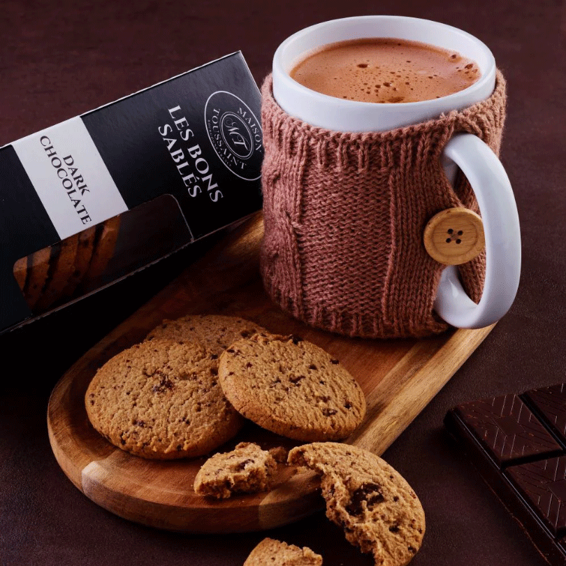 Boîte de sablés Les Bons Sablés au chocolat noir de la Maison Toussaint, accompagnée de cookies aux pépites de chocolat posés sur une planche en bois, et d'une tasse de chocolat chaud dans un cache-mug tricoté avec un bouton en bois. 