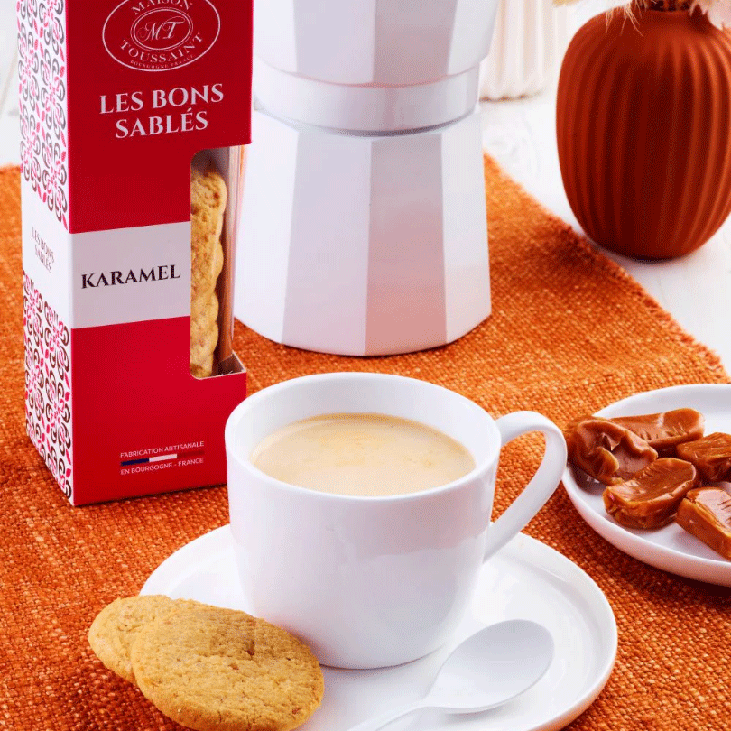 Boîte de sablés Les Bons Sablés au caramel de la Maison Toussaint, posée à côté d'une tasse de café crème sur une nappe orange.