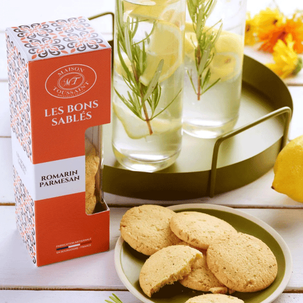 Boîte de sablés Les Bons Sablés au romarin et parmesan de la Maison Toussaint, présentée avec des biscuits sur une assiette verte. Deux verres de citronnade avec des branches de romarin sont placés à côté sur un plateau rond doré, avec des citrons en arrière-plan.