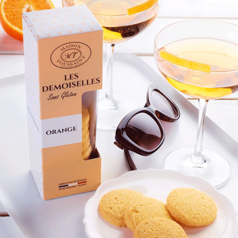 Boîte de biscuits Les Demoiselles à l'orange de la Maison Toussaint, disposée sur un plateau avec des lunettes de soleil et deux verres de cocktail garnis de zestes d'orange. 