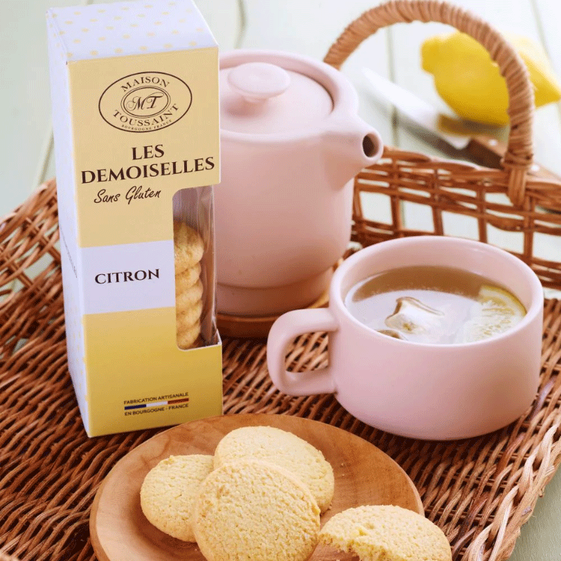 Boîte de biscuits Les Demoiselles au citron de la Maison Toussaint, posée dans un panier en osier avec une tasse de thé au citron et une théière rose en arrière-plan.