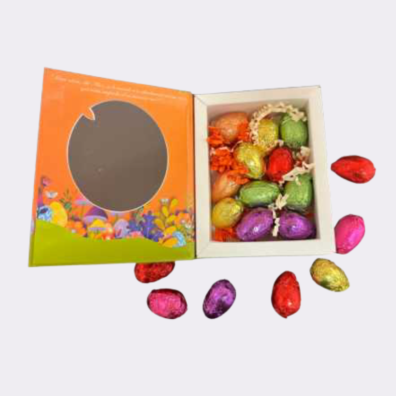 Boîte ouverte de chocolats pralinés "Les Histoires du Lapin d’Alice" contenant 11 œufs en chocolat emballés dans des papiers colorés brillants (rouge, vert, doré, violet, rose), disposés dans un écrin avec des décorations en papier orange et blanc. Le coffret est décoré avec des motifs printaniers et de Pâques, avec des fleurs et des œufs colorés.