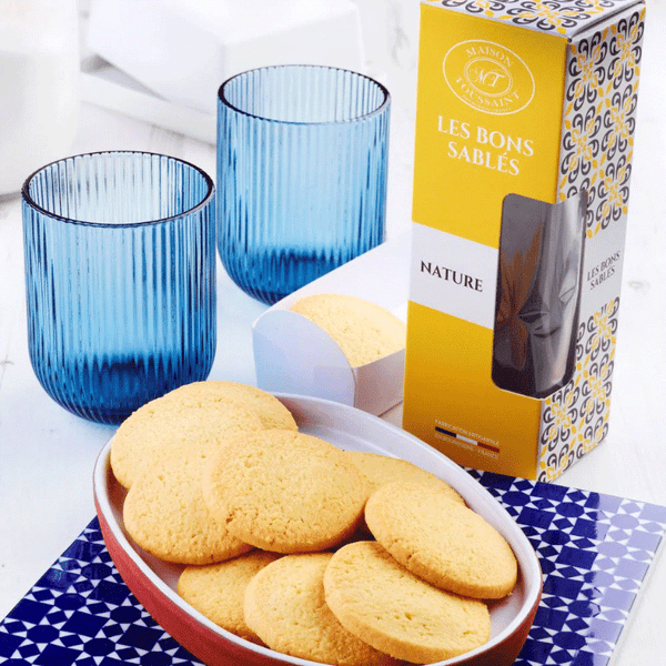 Sablés nature artisanaux présentés en coupelle, avec étui Maison Toussaint et verres bleus sur table lumineuse.