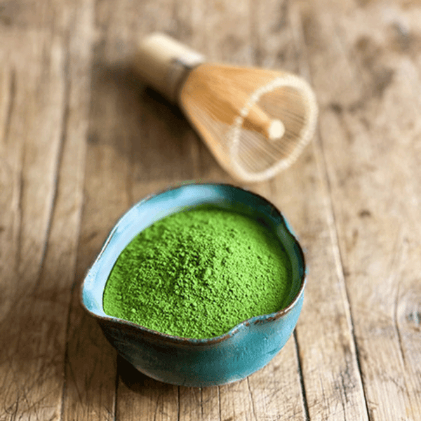Bol en céramique bleue rempli de poudre de thé matcha d’un vert éclatant, posé sur une table en bois rustique. En arrière-plan, un fouet traditionnel en bambou (chasen) utilisé pour la préparation du matcha. L’ensemble évoque une ambiance artisanale et japonaise, centrée sur la cérémonie du thé.