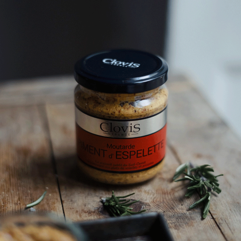 Pot de moutarde au piment d'Espelette Clovis France posé sur une table en bois rustique. Mise en scène chaleureuse avec des branches d’herbes aromatiques.