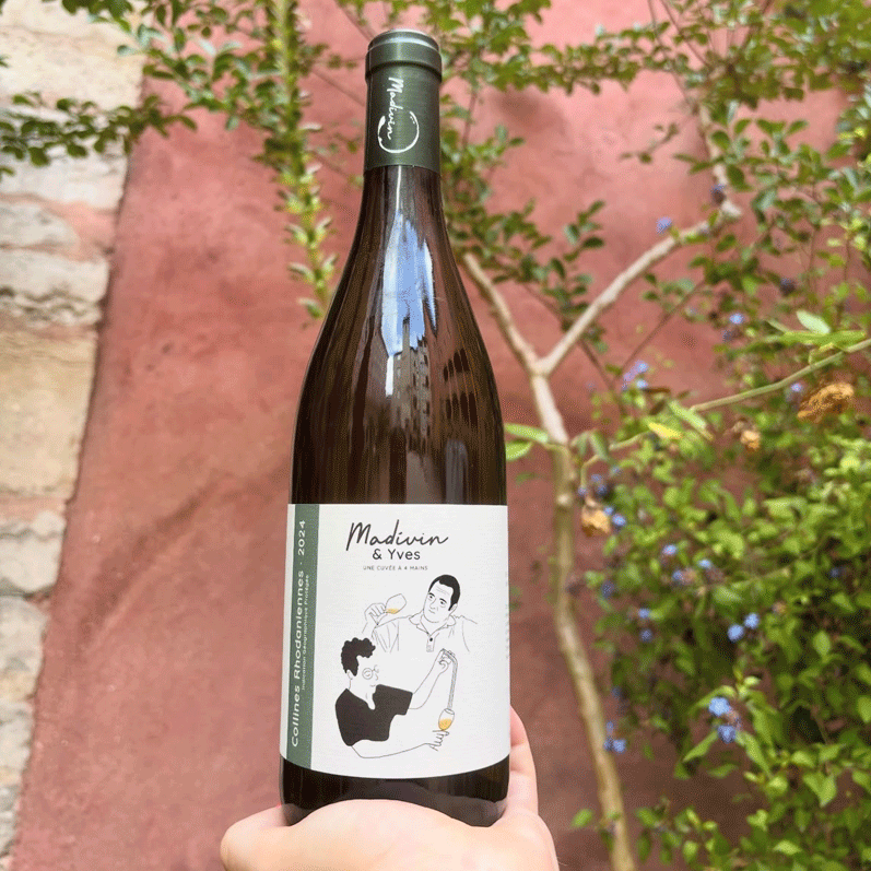 Bouteille de vin “Madivin & Yves 2024” tenue à la main, fond rose et feuillage.