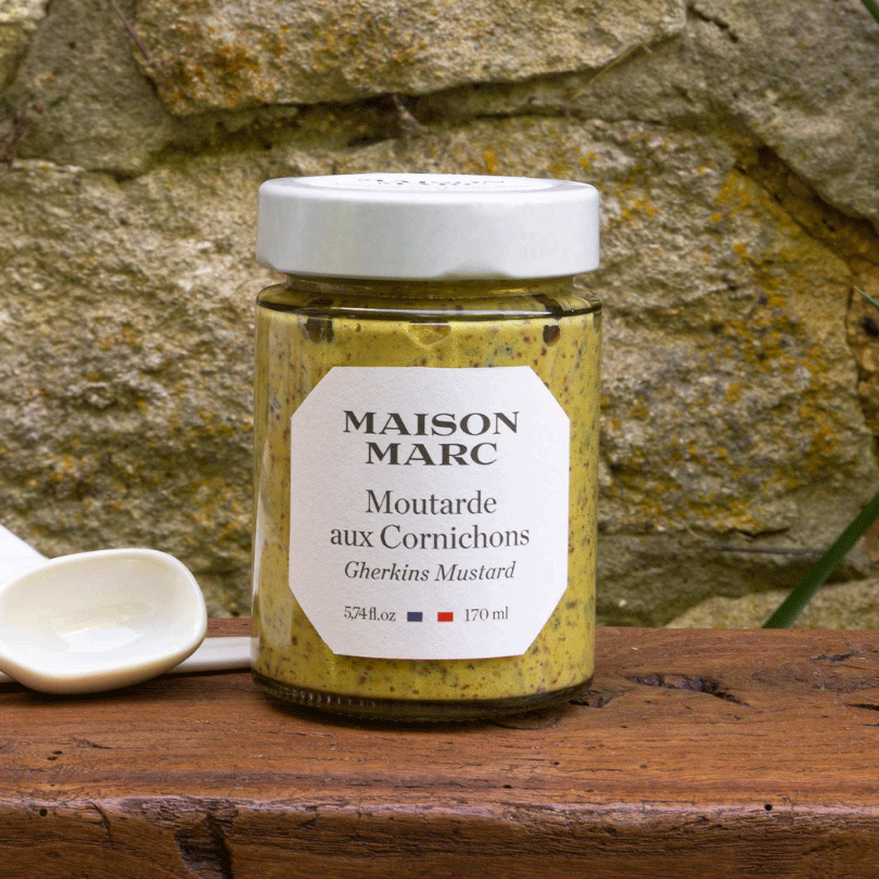 Bocal de moutarde aux cornichons Maison Marc, texture granuleuse visible, posé devant un mur en pierre.