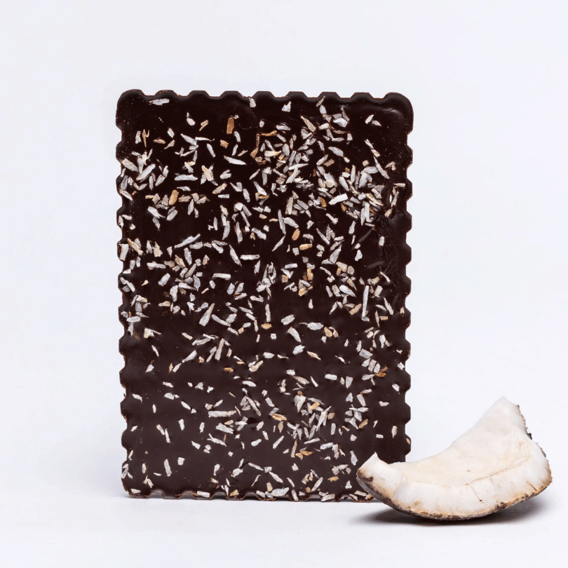 Tablette de chocolat noir 75% bio à la noix de coco, décorée de copeaux de coco grillés, accompagnée d'un morceau de noix de coco fraîche.