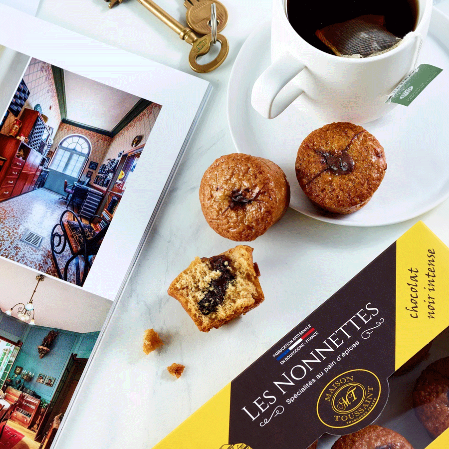 Scène de petit-déjeuner avec une boîte de nonnettes au chocolat noir intense de la Maison Toussaint, accompagnée de nonnettes coupées révélant une garniture chocolatée fondante, une tasse de thé chaud et un livre ouvert avec des clés dorées posées à côté.