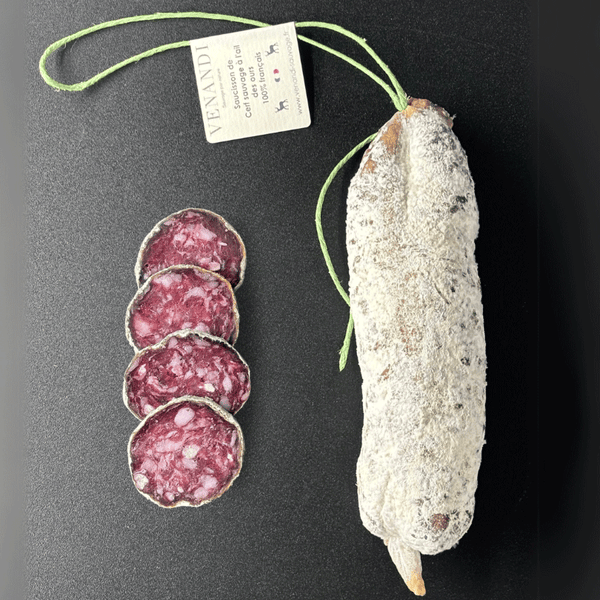 Un saucisson au cerf sauvage 100 % français, parfumé à l’ail des ours, repose sur un fond noir élégant. L’étiquette artisanale de la maison Venandi, attachée par une ficelle verte.