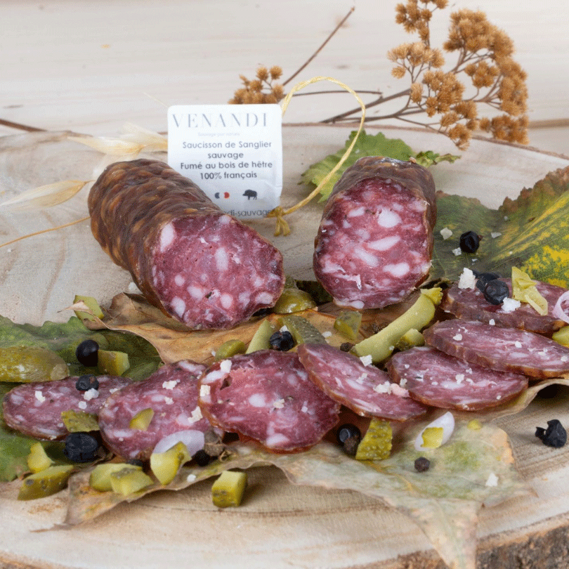Le saucisson de sanglier sauvage, fumé au bois de hêtre, est mis en scène de façon gourmande sur une planche en bois. Tranché généreusement, il est entouré de morceaux de cornichons.