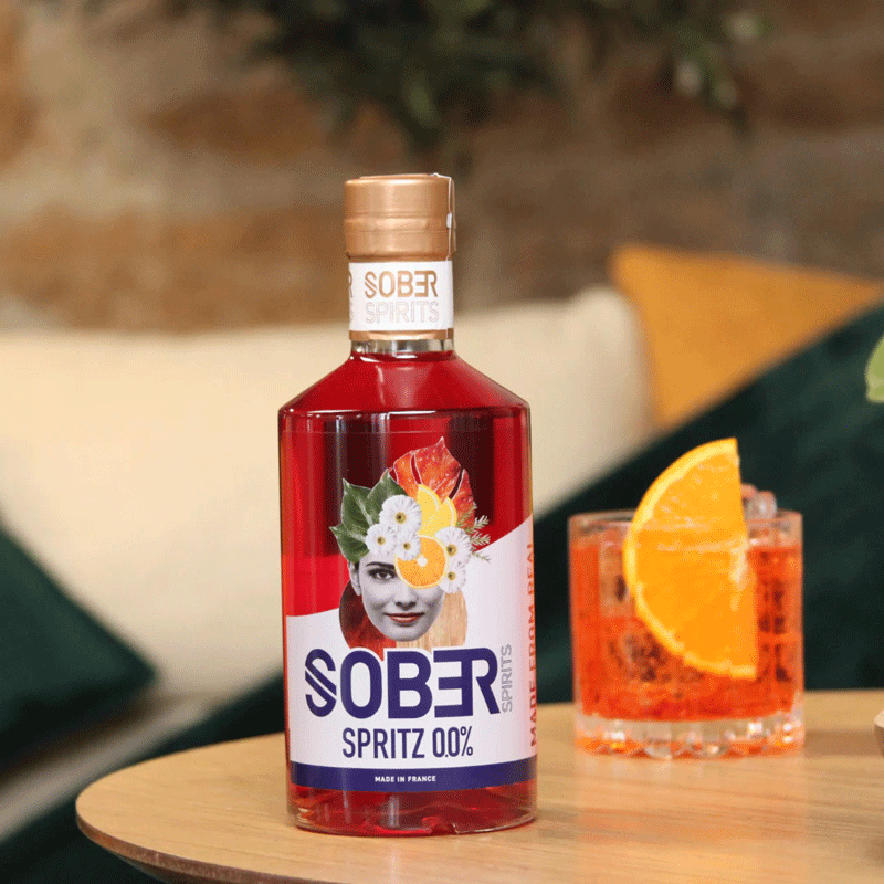 Bouteille de Spritz sans alcool Sober Spirits sur table, fond cosy avec verre garni d’une tranche d’orange.