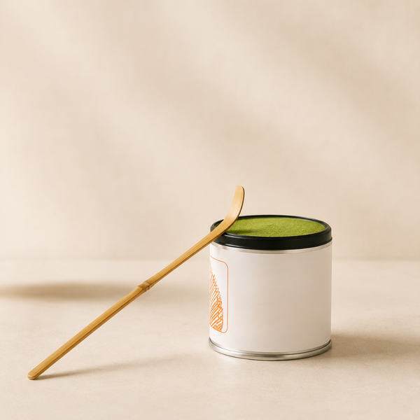 Boîte de thé matcha en métal avec poudre verte visible, accompagnée d’une spatule en bambou sur fond clair.