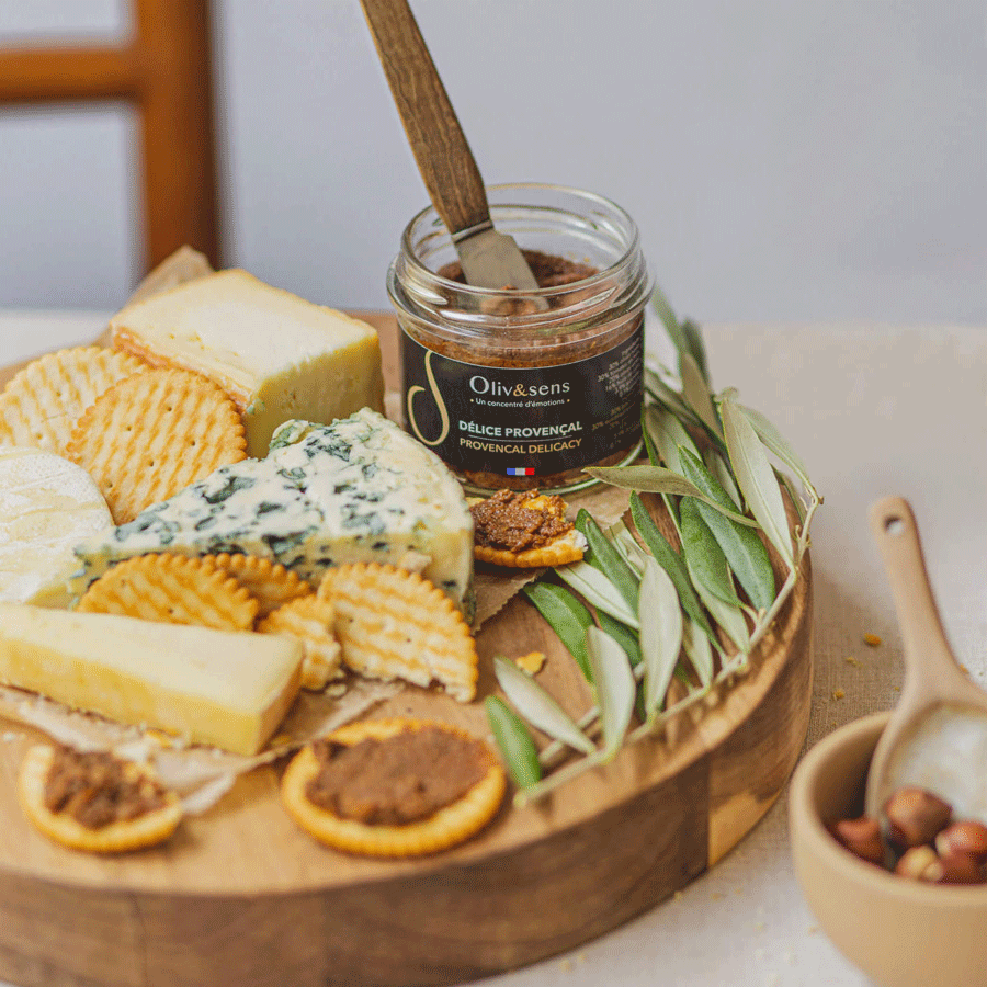 Pot ouvert de « Délice Provençal » Oliv&sens sur une planche en bois entouré de fromages variés, de crackers tartinés et de branches d’olivier. 
