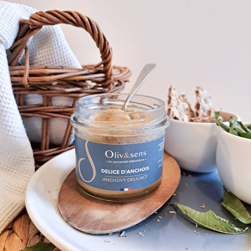 Tartinade Délice d’anchois Oliv&sens, pot de 90 g. Un petit pot en verre contenant une crème d’anchois onctueuse beige, avec une cuillère plongée à l’intérieur. L’étiquette bleu marine affiche la marque et le nom du produit en français et en anglais. Présenté sur une planche en bois, accompagné de pain croustillant et de feuilles fraîches, dans une ambiance apéritive et artisanale.