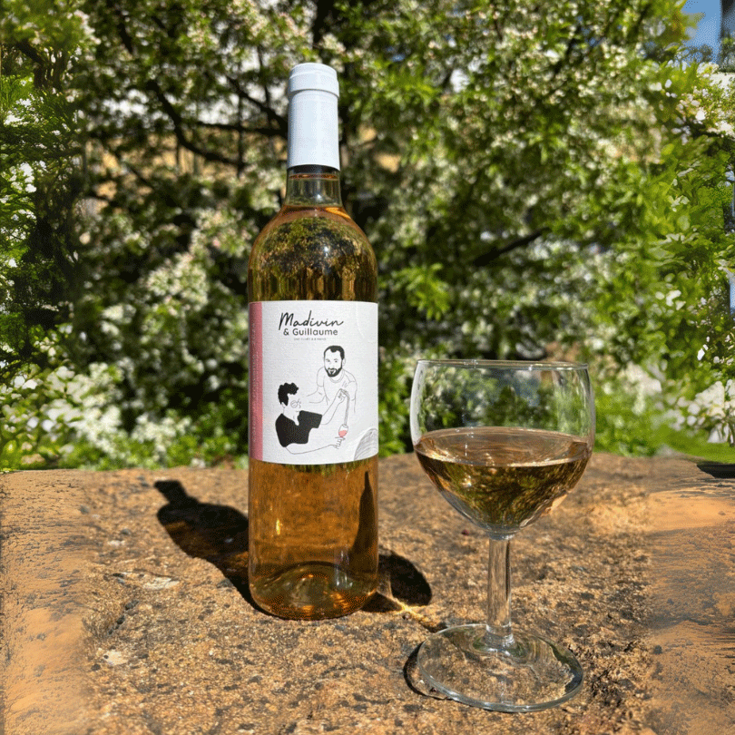 Bouteille de vin rosé « Madivin & Guillaume » Côtes de Provence 2024, posée sur un muret en pierre, accompagnée d’un verre rempli du vin. En arrière-plan, un jardin verdoyant et fleuri baigné de lumière printanière.