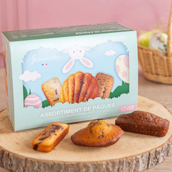 Boîte d’assortiment de Pâques au beurre d’Isigny AOP de la biscuiterie Jeannette, posée sur une planche en bois avec trois madeleines variées devant. En arrière-plan, un panier avec des œufs.