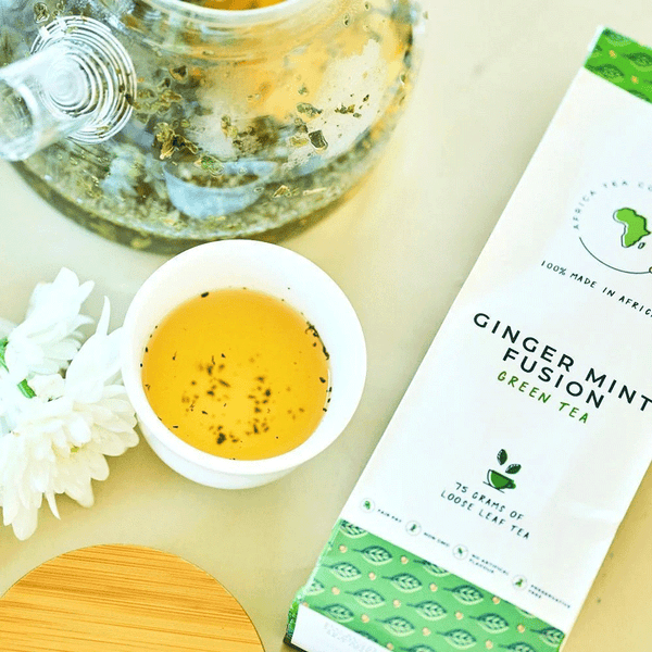 Tasse de thé infusé avec sachet Ginger Mint Fusion, théière transparente et fleurs blanches.