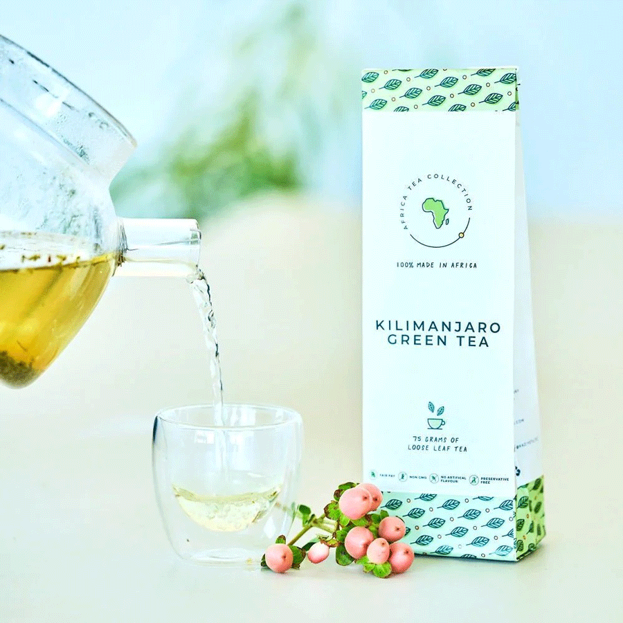 Théière en verre versant du thé vert dans une tasse, avec sachet Kilimanjaro Green Tea et baies roses.