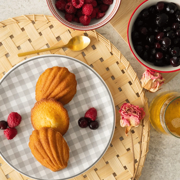 Assiette à carreaux avec trois madeleines saveur framboise, entourée de framboises fraîches, de baies noires et de pétales de rose séchés. Un décor soigné avec une cuillère dorée et des bols de fruits sur un plateau en bambou.