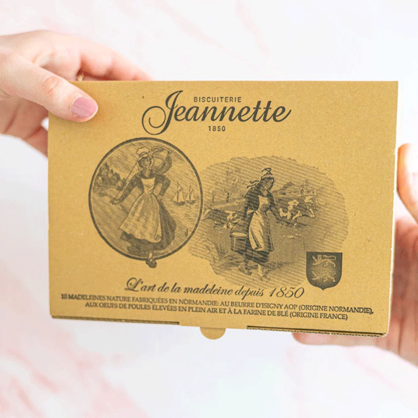 Boîte en carton kraft de madeleines nature de la biscuiterie Jeannette, tenue entre deux mains. Le packaging affiche des illustrations vintage représentant des scènes artisanales et la mention "L’art de la madeleine depuis 1850". 